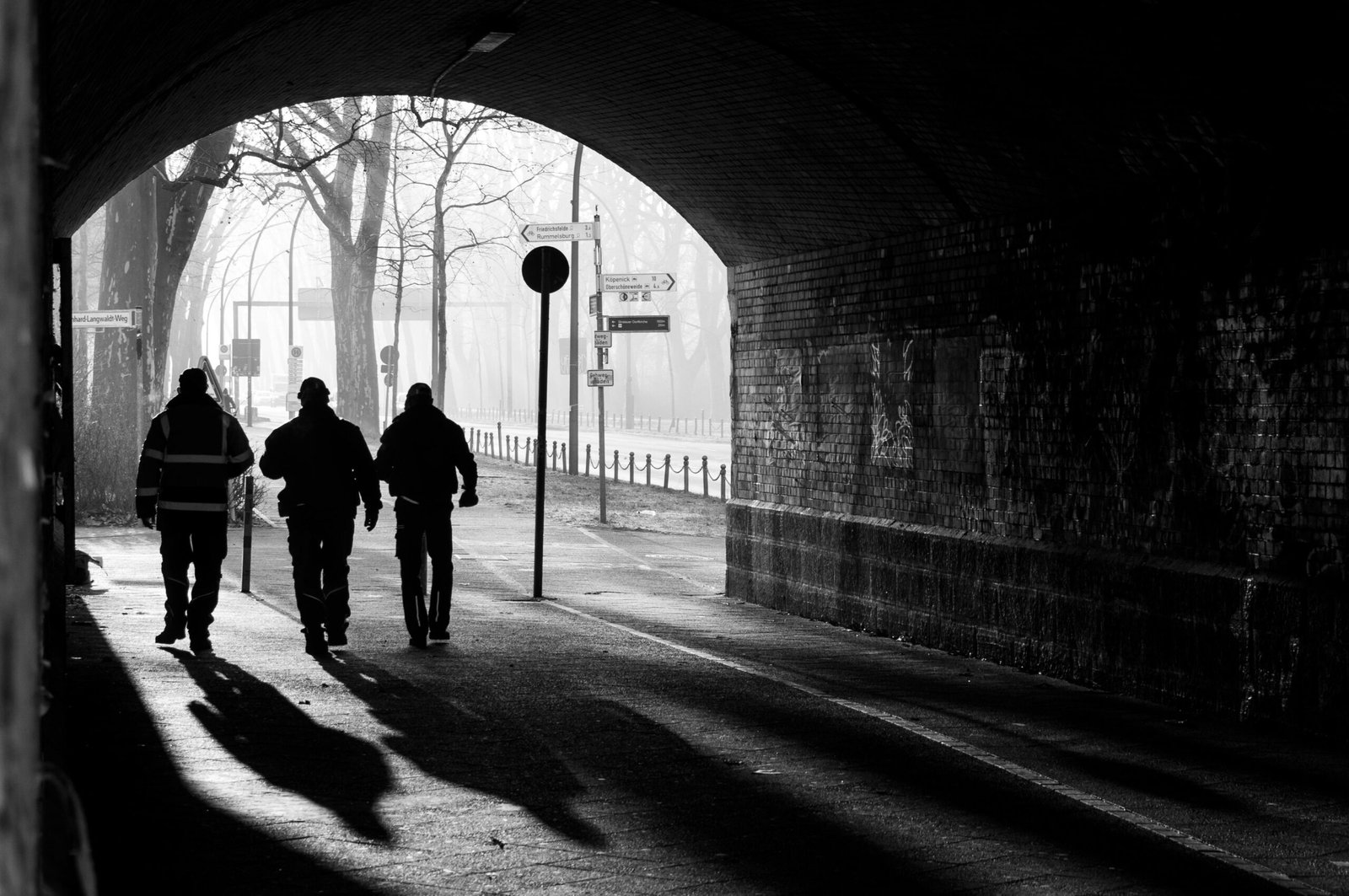 Black and white image of three people walking under a tunnel with dramatic shadows.